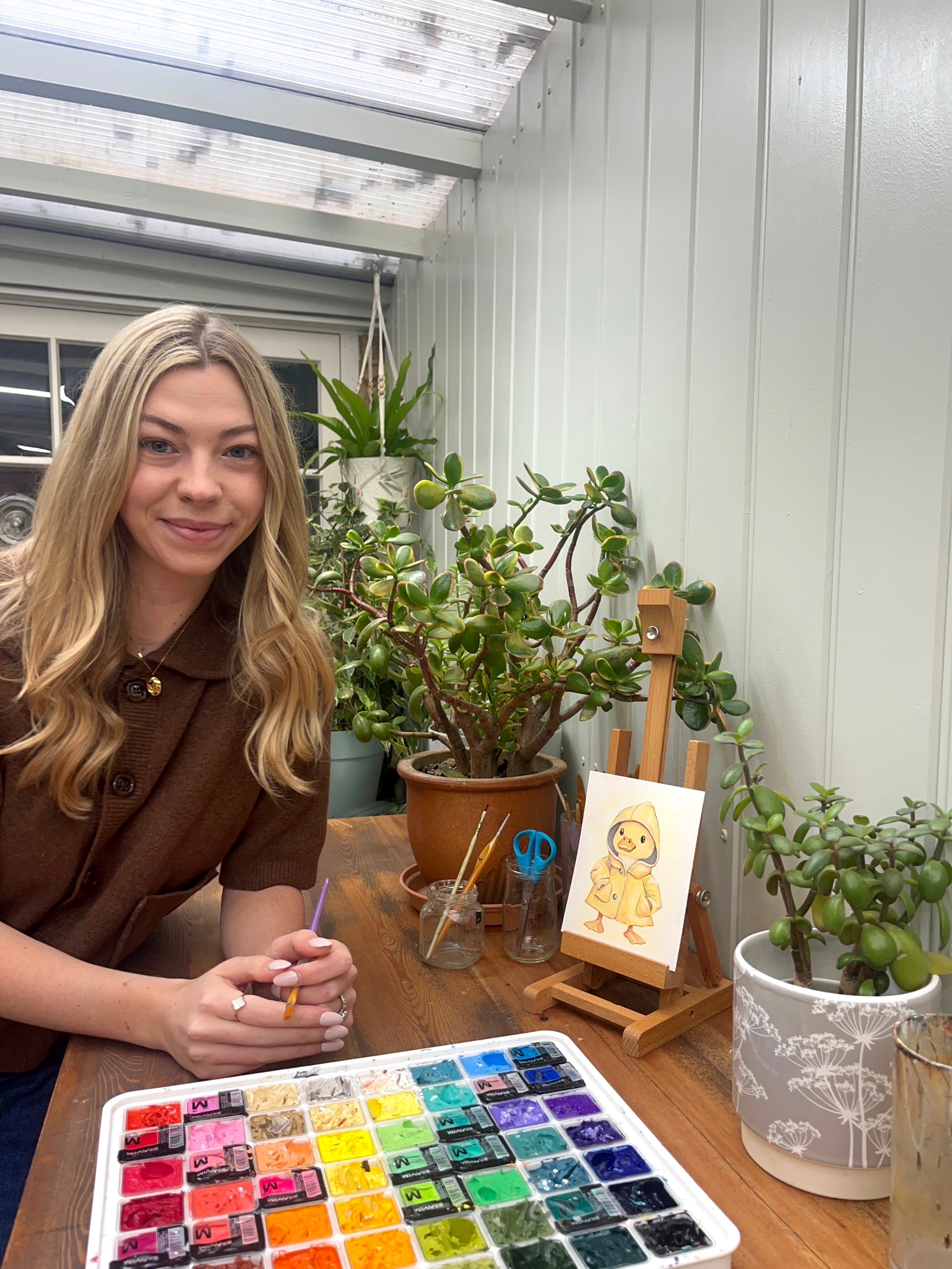 Woman sitting at a table with watercolor paints and plants in a greenhouse.