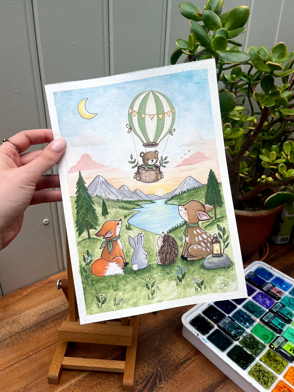 Illustration of animals by a lake with a hot air balloon, held on a wooden easel.