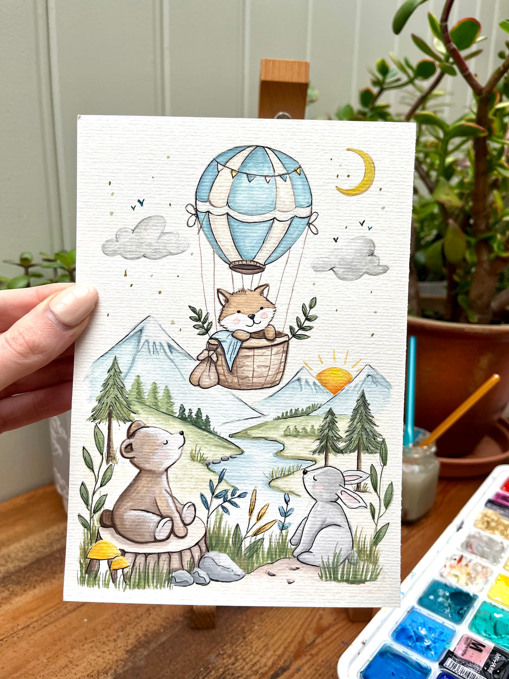 Illustrated card with fox, rabbit, and hot air balloon in a scenic setting held by a hand.