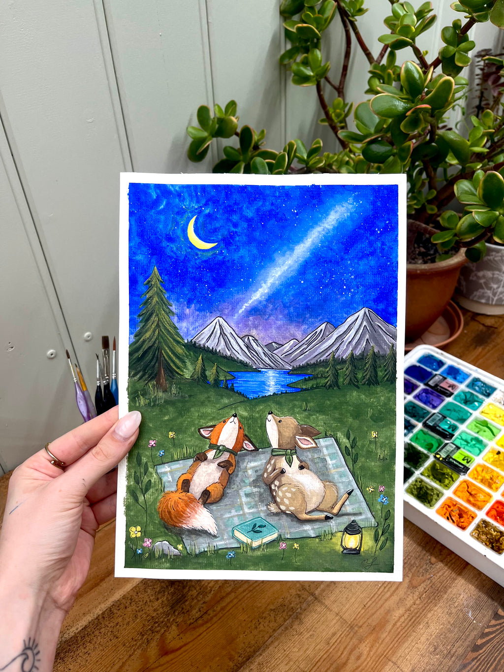Hand holding a painting of animals by a lake with mountains and a starry sky, surrounded by plants and art supplies.