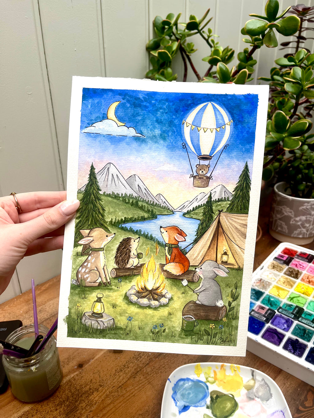 Illustration of animals around a campfire with mountains and a hot air balloon in the background, held by a hand on a wooden surface.