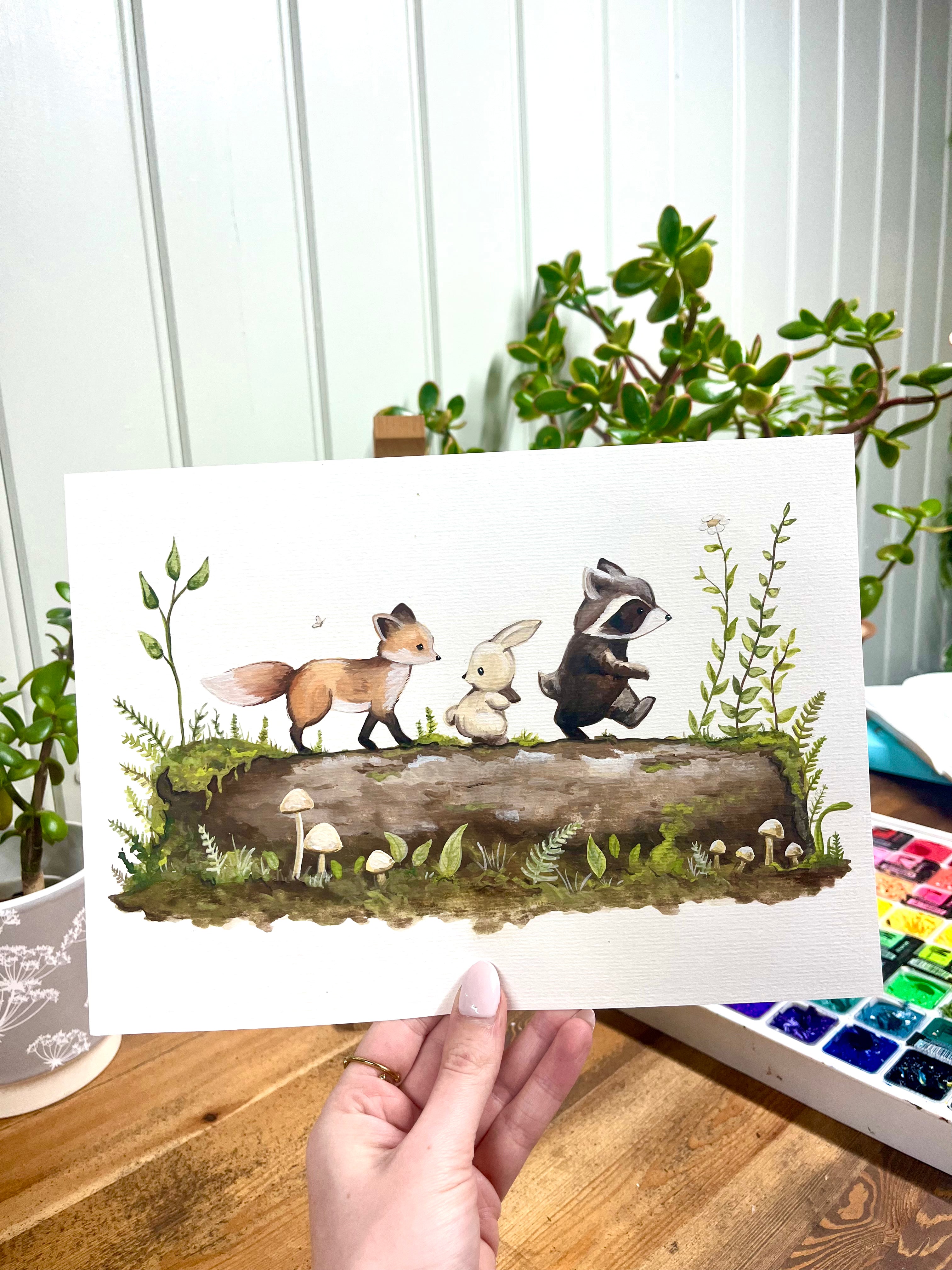 Hand holding a print of a forest scene with animals on a log.